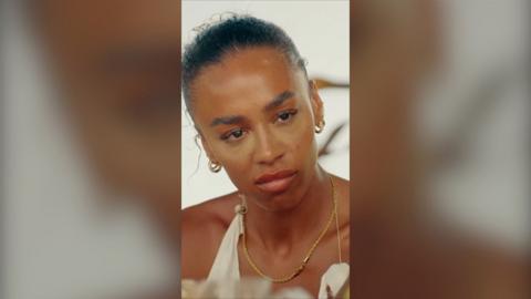 A woman looks annoyed. Her hair is slicked back, she wears a shoulderless white top and gold hoop earrings.