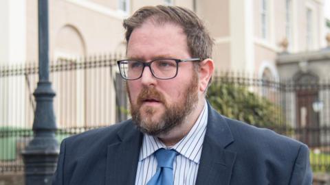 Alan Trevor Campbell outside court, he has short brown hair with grey bits and a beard. He is wearing dark framed glasses, a grey/blue suit with a white shirt with blue pinstripes and a light blue tie. 