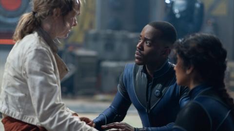 Woman with white jacket sits opposite a man and woman wearing futuristic blue and black jumpsuits.