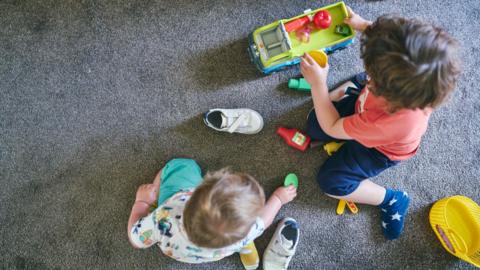 As seen from above, two children are sat on a grey carpet. One has fair hair and is wearing green shorts and a white printed top. Their shoes are next to them and they are playing with a green disc. One has dark hair and is wearing an orange top and navy shorts and socks. They are playing with a truck with plasic food items in it.
