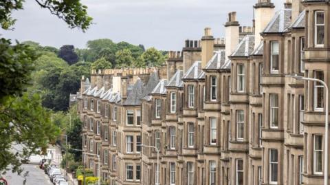 A row of closely packed, beige, stone-terraced houses with bay windows and tall chimneys line a slightly sloping residential street. Parked cars run along the left side of the road, while a leafy tree branch frames the scene in the foreground