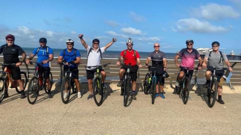 Eight men in cycling gear line up along a seafront while straddling their bikes on a sunny day. They are smiling, with one of the line-up holding their hands up in the air.