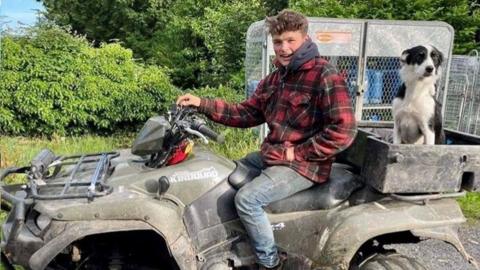 A teenager sitting on a quad bike. The teenager is smiling at the camera and has placed one hand on the handle bars and the other is in his coat pocket. A dog is sitting on the back of the quad bike. Bushes are in the background. 