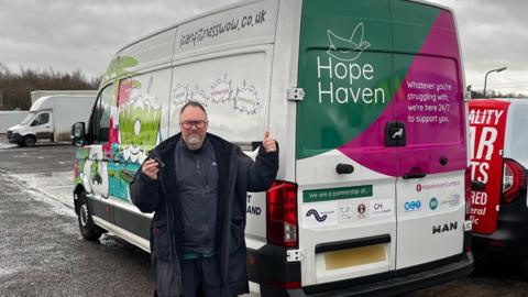 Phil Stout holding a set of keys and giving a thumbs-up gesture as he stands in front of the "wheels of wellness" van. He has short dark hair, a grey beard and glasses. The white van is decorated with the charity's name and colourful graphics.