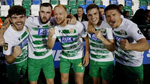 Five football players in green shorts and white shirts with green stripes clench fists in victory while smiling at the photographer.