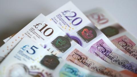 Four bank notes spread out on a white table. The notes are a £5, £10, £20 and £50 note.