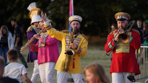 Three men playing brass instruments in a park, wearing colourful army style jackets and white pants.