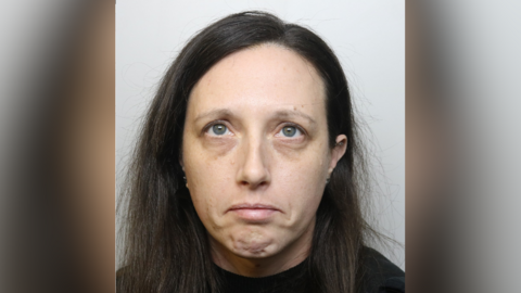 A police custody image of Heidi Bessant. She has shoulder length dark hair and wears a dark top. She is looking to the ceiling and frowning. 