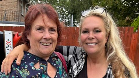 Karen Strong and Samantha Southern standing in a garden. Samantha has long blonde hair and glasses on the top of her head, and is standing with her right arm round Karen's shoulders. She has red hair and both are smiling