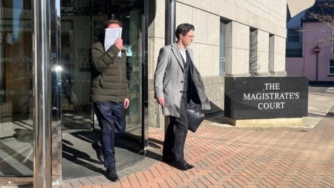 Two men leaving Jersey's Magistrate's Court. On the left, a man is covering his face with a white envelope. He is wearing a dark green puffa jacket and blue trousers. On the right, a man is wearing a light grey coat and black trousers. He is carrying a black briefcase. A sign, saying 'The Magistrate's Court', is visible to the right of the image.