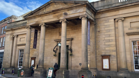 The image shows the front of a historic sandstone building in York. The architecture is classical in style. The building’s façade is dominated by a portico with six tall stone columns supporting a triangular pediment. The columns have decorative capitals and stand evenly spaced across the front. The stonework is light beige with visible weathering and darker patches.