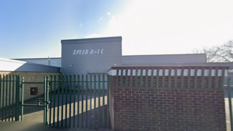 A grey building with a white sign reading Speedball and a car park in front and a partial brick wall and fences