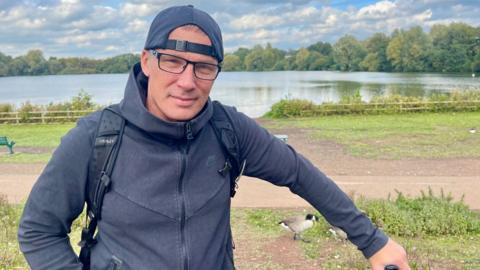 A man in stands beside a lake. He is wearing a dark zipped hoodie, a black rucksack, glasses and a dark baseball cap worn backwards. His left hand is holding the handlebar of a bicycle. Geese can be seen wandering in the background beside a footpath.