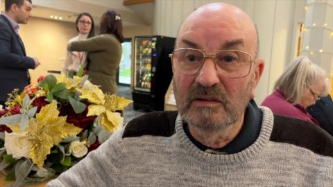 A bald man in glasses with a short grey and brown beard, wearing a grey sweater with brown shoulder coverings, looks slightly to the left of camera. Behind him over his right shoulder are three members of staff, a man and two women,  in conversation, with a flowery table decoration in the foreground. Behind the man's left shoulder is a seated woman looking down. She has grey hair in a pony tail.  
