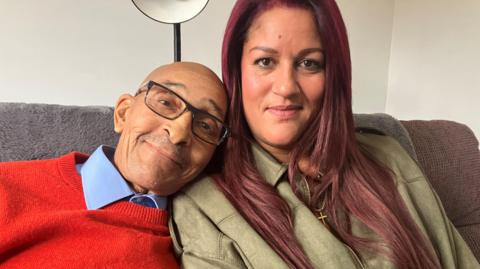 A man wearing a red jumper and blue shirt and wearing glasses. His head on his daughter's shoulder. She is wearing a light green coat and has brown/burgundy hair and is wearing a gold cross. 