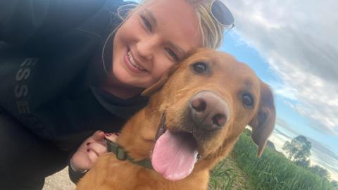 Sophie Theakston has blonde hair and blue eyes. She is wearing sunglasses on top of her head and a black hooded sweatshirt. She is hugging her pet Bertie while holding onto his collar. Bertie is a red-coloured Labrador and has his tongue hanging out. Both are looking at the camera.