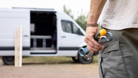 A close-up of a person's hand holding tools. They are wearing a white shirt and grey trousers and are standing in front of a white van with its door open and wooden planks leaning on the side of the vehicle.