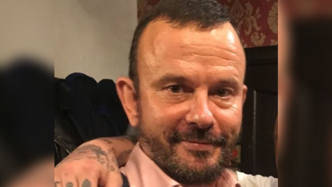 A head and shoulder photo of Eddie Roberts looking into the camera. He has a dark beard and short dark hair. He is wearing a pink shirt and has someone's arm over his shoulder. 