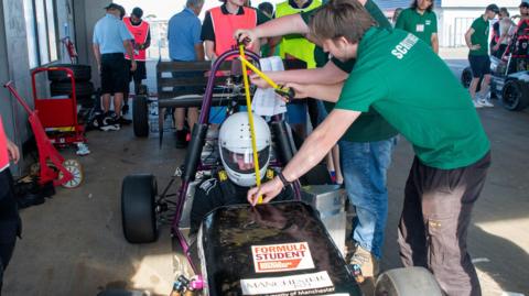 Men measuring parts of a racing car with a driver seated inside wearing a white helmet