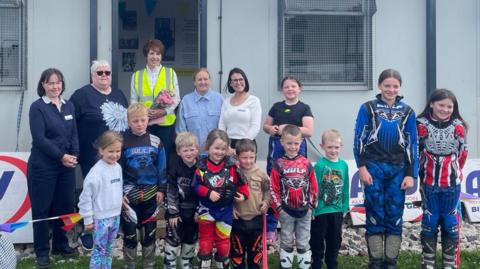 Ten children, some of whom wearing colourful motorbike gear, smile for the cameras. Five adults stand behind them in front of a grey hut.