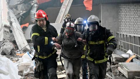 Three firefighters in full gear and oxygen masks walk out a woman in a face mask after Russian strike on residential building in Kharkiv