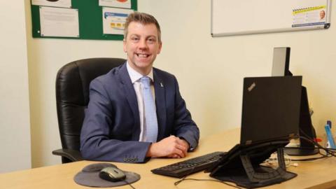 A middle aged man with short brown hair who is wearing a navy blazer, white shirt, and light blue tie smiles as he sits in front of his computer in an office, his hands folded on the desk in front of him.