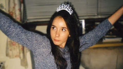 A picture of Fiona Braidwood wearing a long sleeved grey top and a sparkly tiara on her long dark hair. She is sitting in a living room in front of a window, lifting both her arms high and looking off to the left of the camera.