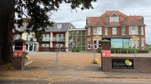 A building and the front of the hotel can be seen. There is a brick wall with a sign saying 'The Stanwell Hotel' and fencing surround the hotel. The sky is grey.