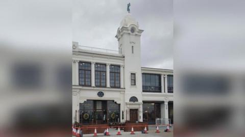 The Split Chimp which has a dark brown wood shop front cordoned off by orange and white cones. The shop is part of the white Spanish City building. The green dancing lady statue can be seen directly above the bar on top of a domed tower.