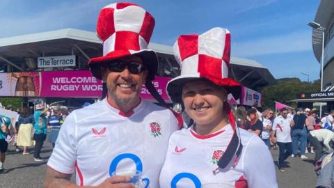 A man and a woman in white England rugby shirts. They are also wearing red and white hats.