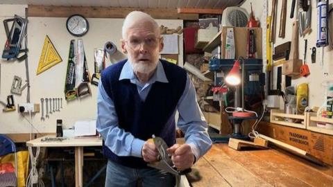 Bruce Foster, wearing a light blue shirt, dark blue knitted tank top and narrow jeans, inside the Men's Shed. He is using a planing tool on wood, secured in a vice, attached to a wooden bench. 