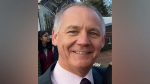 Paul Taylor in a blazer, white shirt and tie smiling at the camera. 