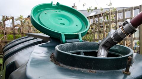 A large plastic heating oil tank in a garden with its top open and a hose filling it.