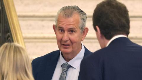 A man with short grey hair, wearing a navy suit, blue shirt and silver tie. He is speaking to two people, who are facing with their backs to the camera, also in formal wear. They are stood beside an ornate stone staircase at Stormont.