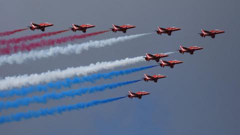 The Red Arrows in flight on a bright sunny day. There are 10 jets in the shape of an arrow. Three have blue vapour trails coming from the aircraft's tails, three others have white and three more have red.