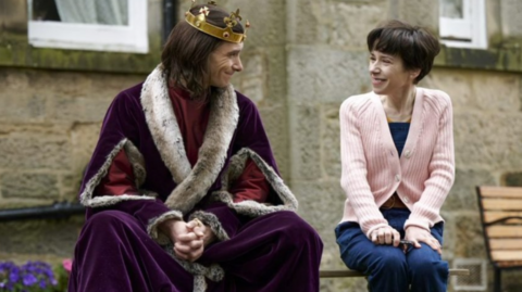 Sally Hawkins, wearing a pink cardigan and jeans, sitting next to an actor in a royal cloak and crown.