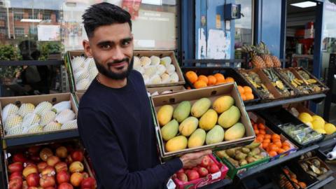 Shopkeeper Shivam Pabari, a young Indian man wearing a long-sleeved sweater carrying an open box of mangoes. He is standing outside a grocery shop in front of a fresh fruit and vegetable display