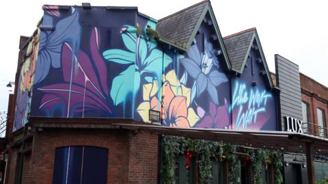 A red brick building which has a dark blue flower painting beneath the roof. There are Christmas garlands hanging down.