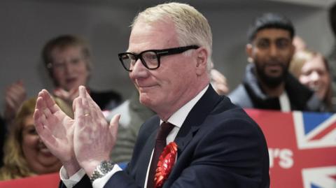 Richard Parker applauds supporters at his election count in May. He is wearing a black suit, and is surrounded by supporters holding flags and banners.