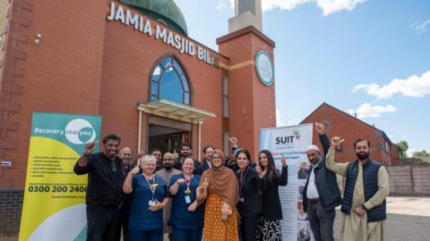 A group of people stand outside a brick building with their hands in the air. Posters can be seen behind them.