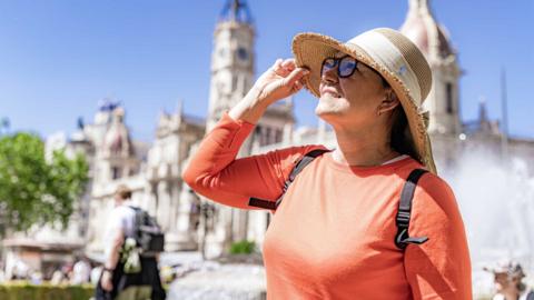 An older woman in a sunny city wearing an orange top, hat and sunglasses