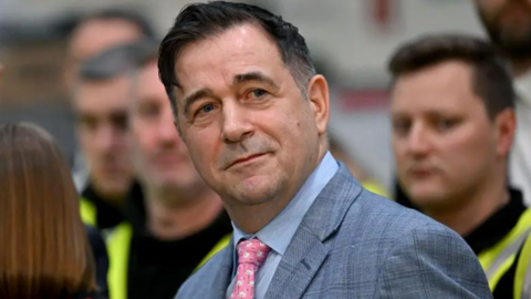 Juergen Maier wearing a grey suit and blue-grey shirt with a pink tie. He has dark hair and is looking slightly off camera. Blurred behind him are three people wearing hi-viz vests in luminous yellow over dark clothing.