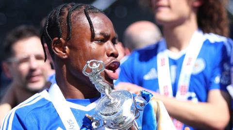 Abdul Fatawu, wearing Leicester's blue home kit, lifts the silver Championship trophy with sweat dripping down his face with his team mates in the background behind him