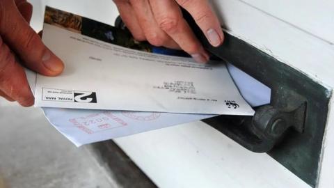 File photo of hand putting letters into a black letter box that is close to ground level.