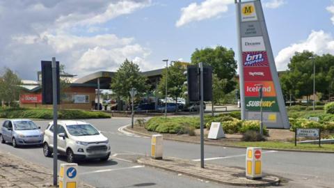 Road entrance to The Speke Centre showing a large sign with names of shops including Morrisons, Dreams, B&M, TK Maxx, Iceland and Subway. A road sign says Penketh Drive and two cars are waiting at a set of traffic lights.