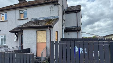 A grey end-terrace house with a boarded up door and windows. There is a grey fence in front of the house and a cloudy sky above. 