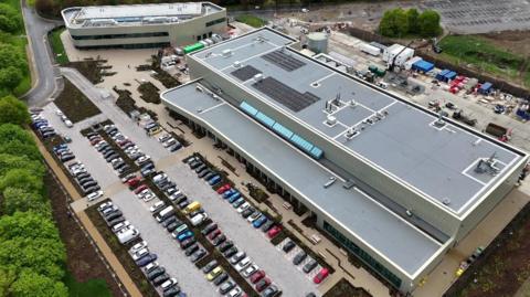 A drone shot of a large, long, modern-looking building, and a smaller one next to it. There is a car park with about 100 cars in it.