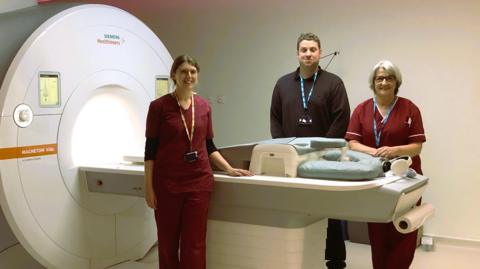 Three members of staff, two in medical clothes, standing beside the donated MRI scanner. It has a tunnel to the left, with a bed to the right of it.