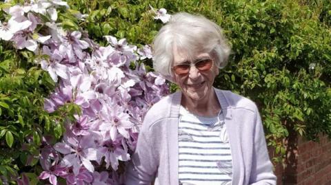 A woman with short white hair, wearing tinted orange glasses, smiles while next to a bush of purples and white flowers, while wearing a lavender cardigan over a white and black striped top.