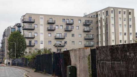 A tall apartment building with grey exterior cladding by a fence and road